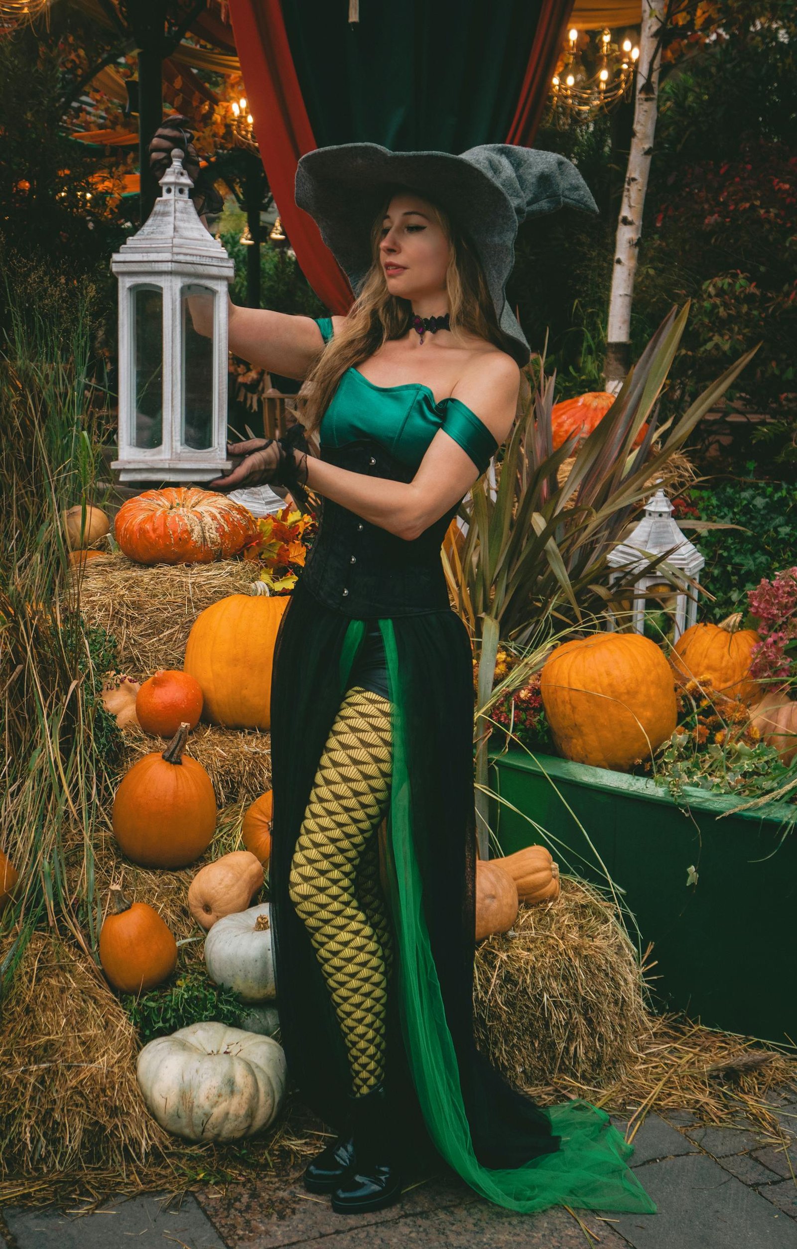 A woman in a witch costume stands by pumpkins outdoors, evoking a Halloween spirit.